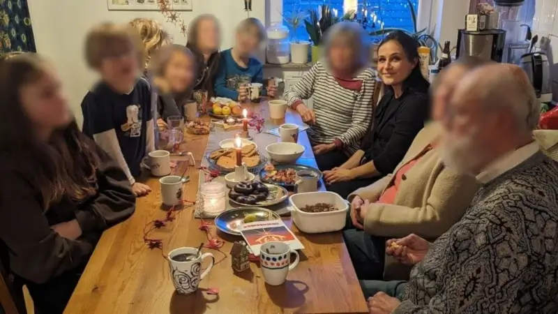 Mehrere Generationen einer Familie sitzen an einem langen Holztisch in einer gemütlichen Küche, trinken Tee und Kaffee, teilen Gebäck und Kerzenlicht in ruhiger Abendstimmung.
