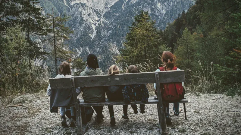 Fünf Kinder und eine erwachsene Person sitzen auf einer Bank vor einer Berglandschaft.
